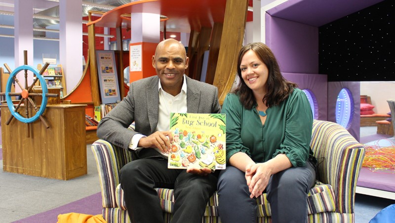 Bristol’s Mayor Marvin Rees (left) and Councillor Ellie King (right), smiling, sit on a sofa in Bristol’s Central Library. Marvin is holding the book ‘First Day at Bug School’. Behind them, books and an open pirate ship are visible to their left. To their right, the sensory nook, including a night sky screen, can be seen.